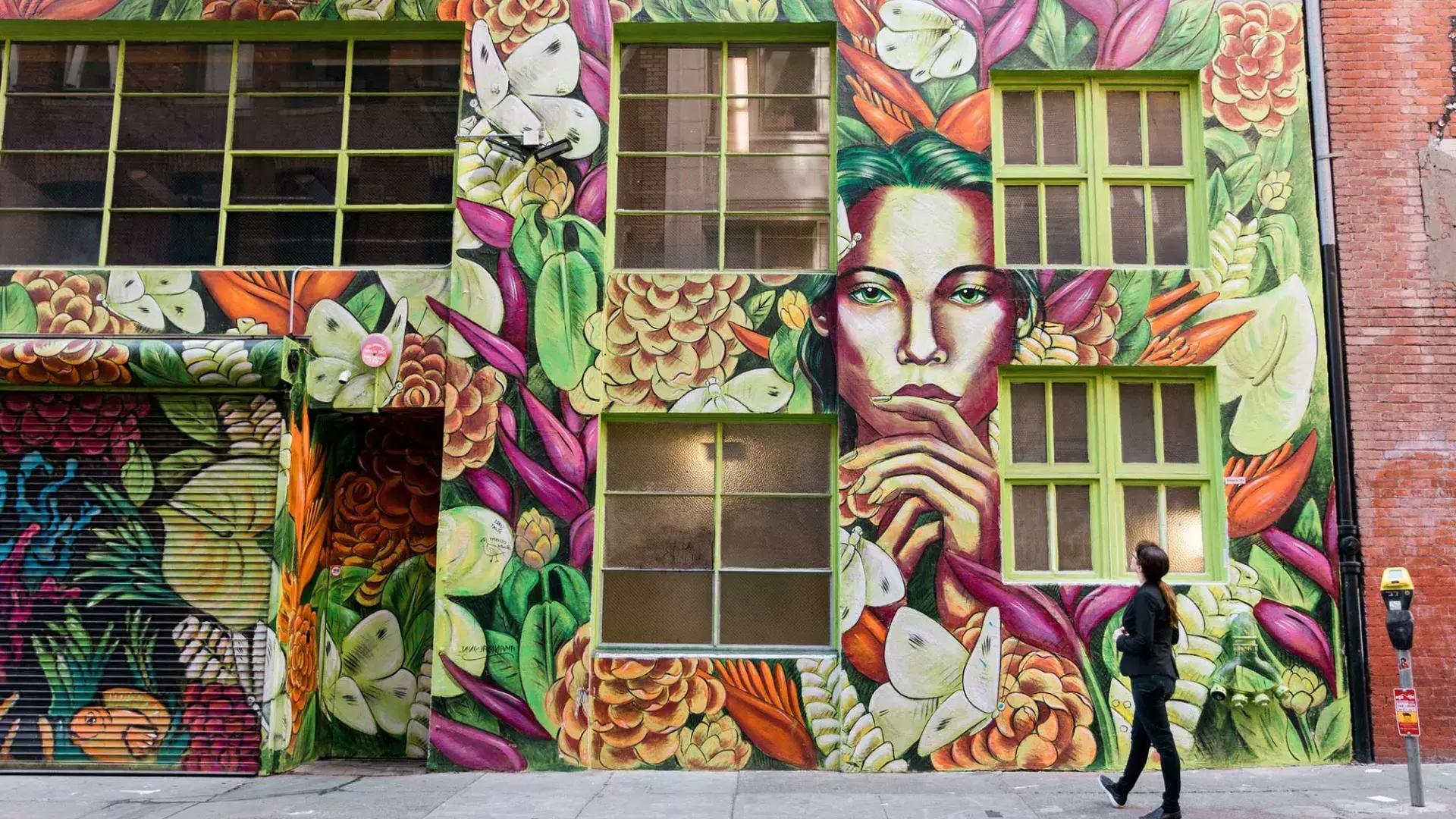 贝博体彩app，一名妇女抬头看着一座砖房侧面色彩鲜艳的壁画.