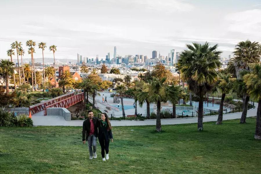 Ein Paar geht auf die Kamera zu, Dolores Park und die Skyline von San Francisco im Rücken.