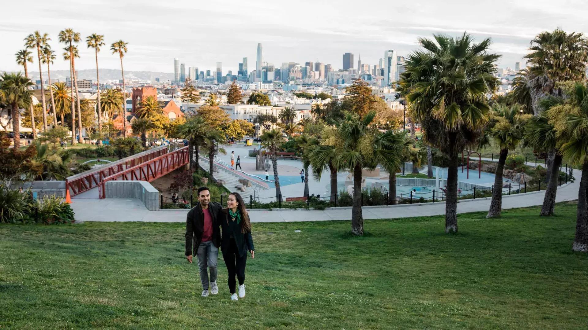 Una pareja camina hacia la cámara con Dolores Park y el Skyline de San Francisco detrás de ellos.
